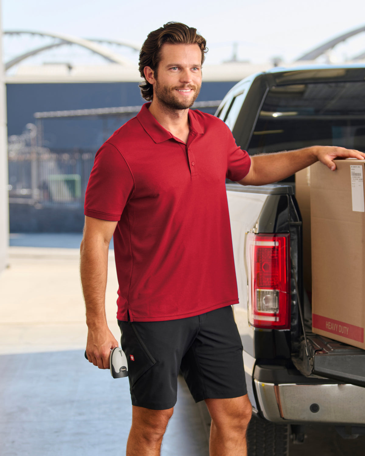 A worker wears the STRAUSS Cotton Stretch Polo Shirt in fire red with tan work pants on a sunny jobsite