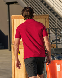 A worker wears the STRAUSS Cotton Stretch Polo Shirt in fire red with tan work pants beside jobsite equipment