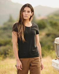 A woman wears the Women's STRAUSS Cotton Stretch T-Shirt in black with work pants on a sunny outdoor workday