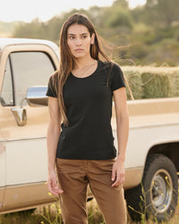 A woman wears the Women's STRAUSS Cotton Stretch T-Shirt in black with work pants on a sunny outdoor workday