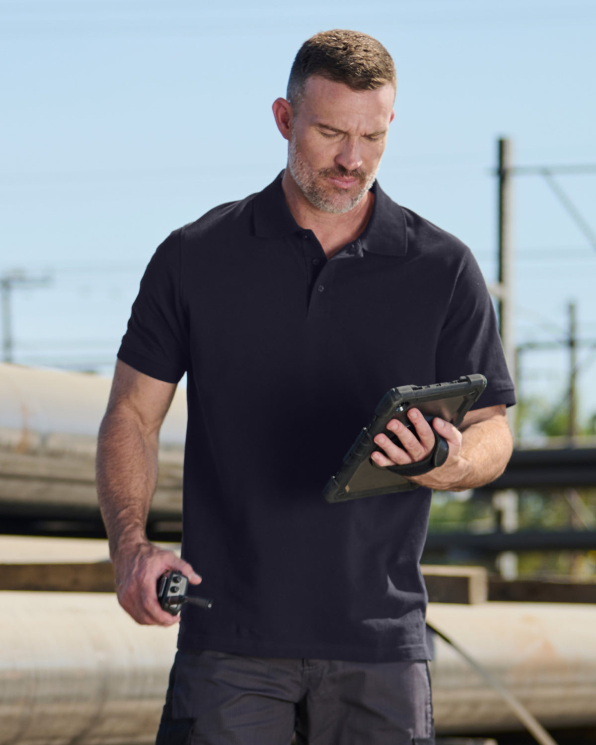 A worker wears the STRAUSS Cotton Polo Shirt in nero blue with tan work pants beside jobsite equipment
