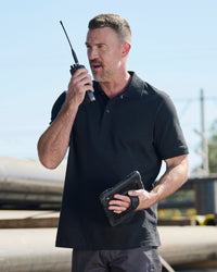 A worker wears the STRAUSS Cotton Polo Shirt in black with tan work pants while working in an industrial setting