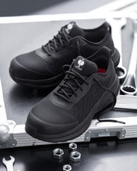 A pair of black work sneakers resting on the corner of a silver metal equipment case, with wrenches and nuts nearby on a dark surface.
