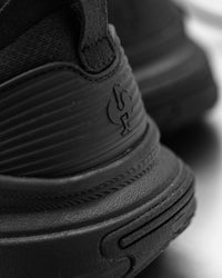 Close-up of a black sneaker showing the textured rubber sole, stitched side panel, and embossed logo.