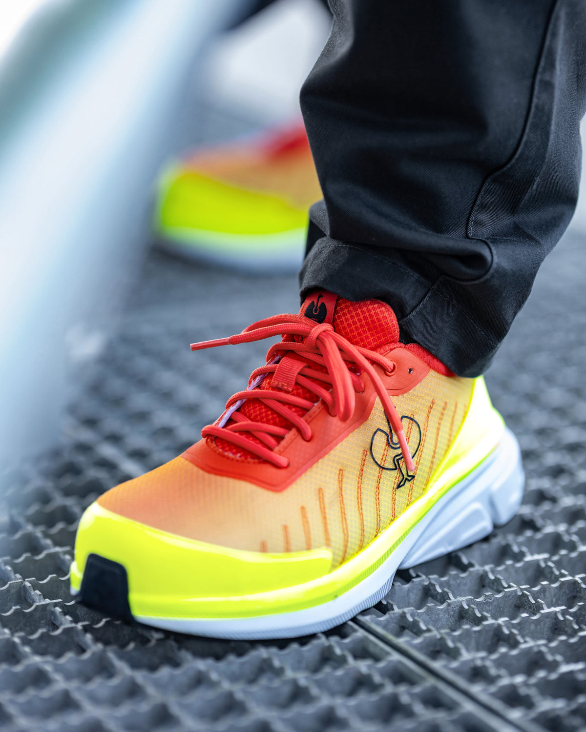 A close-up of a person's foot wearing a vibrant athletic sneaker with a gradient orange-to-yellow upper, neon yellow accents, and red laces, paired with black pants.