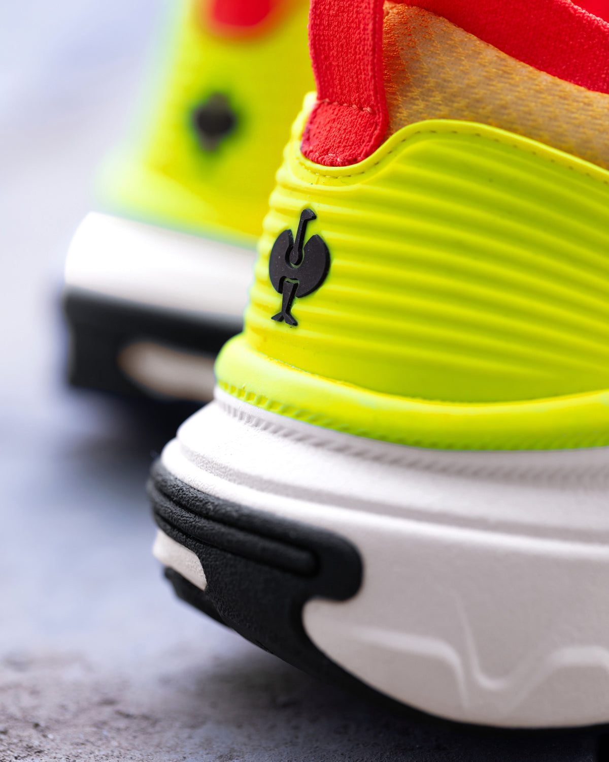 Close-up of a bright neon yellow-green athletic shoe with a red pull tab, white midsole, and black outsole detail, featuring a black rooster logo on the heel.