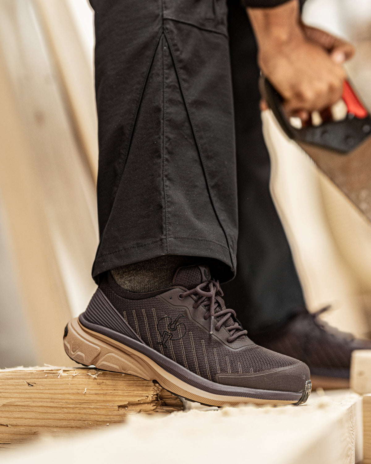 A close-up of a person wearing dark gray and black work boots with a tan sole, standing on a wooden board in a workshop setting.
