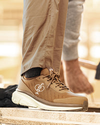 A close-up of a person's foot wearing a tan and brown athletic shoe with a thick white sole, standing on a stone surface. The person is also wearing khaki pants and a gray sweatshirt, with a blurred outdoor background.
