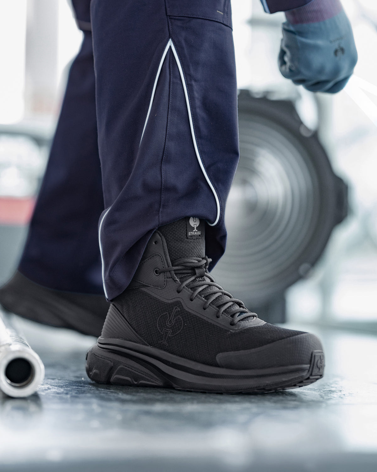 A close-up of a person wearing a dark gray and black high-top work boot with navy work pants featuring white piping, standing on a metallic floor with a blurred industrial background.