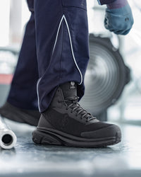 A close-up of a person wearing a dark gray and black high-top work boot with navy work pants featuring white piping, standing on a metallic floor with a blurred industrial background.