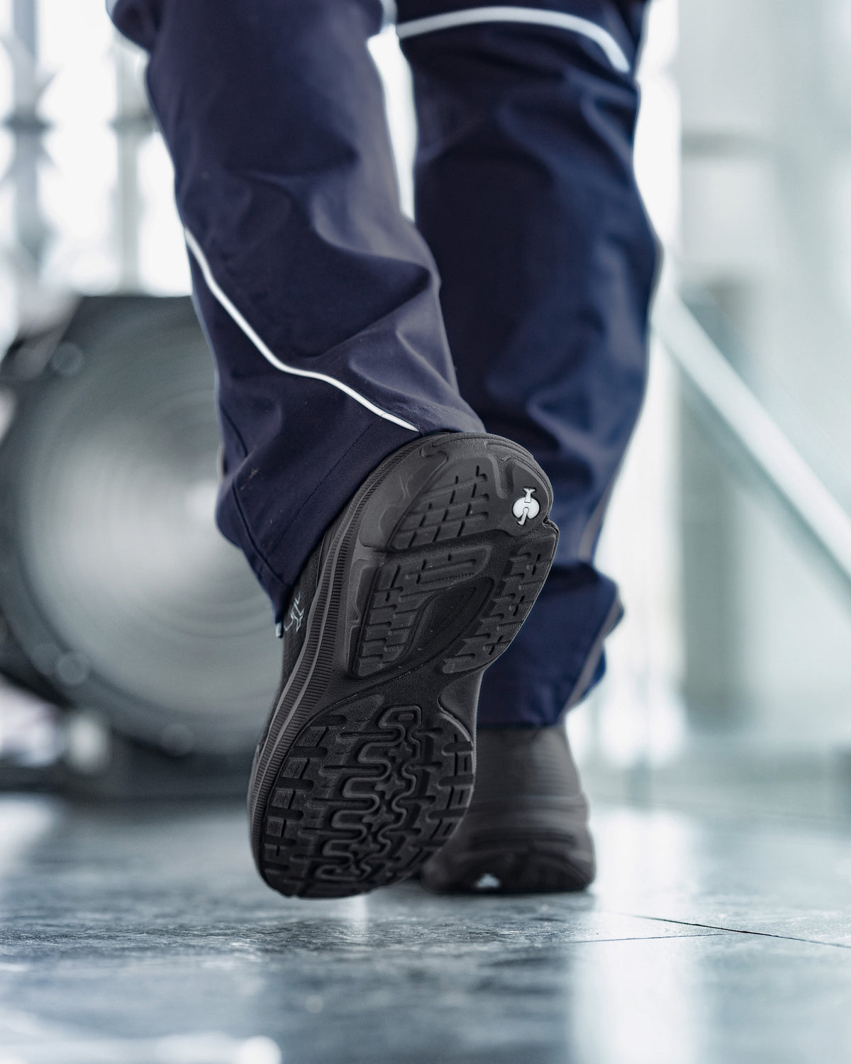 A close-up action shot of a person's legs walking, showing the textured black sole of a work shoe and the side of a navy blue work pants leg with a white stripe.