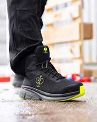 A close-up of a person's foot wearing a black high-top work boot with a bright neon yellow-green logo and sole accent, paired with black pants.