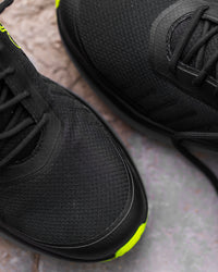 Close-up of a black athletic shoe showing the textured mesh upper, black laces, and a bright neon yellow accent near the sole, set against a gray stone background.