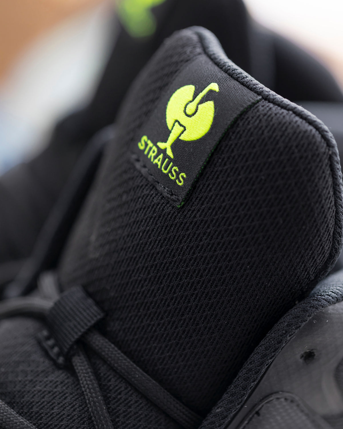 Close-up of a black mesh sneaker showing the tongue label with a bright yellow-green Strauss logo and text.