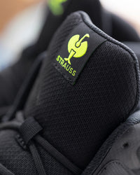 Close-up of a black mesh sneaker showing the tongue label with a bright yellow-green Strauss logo and text.