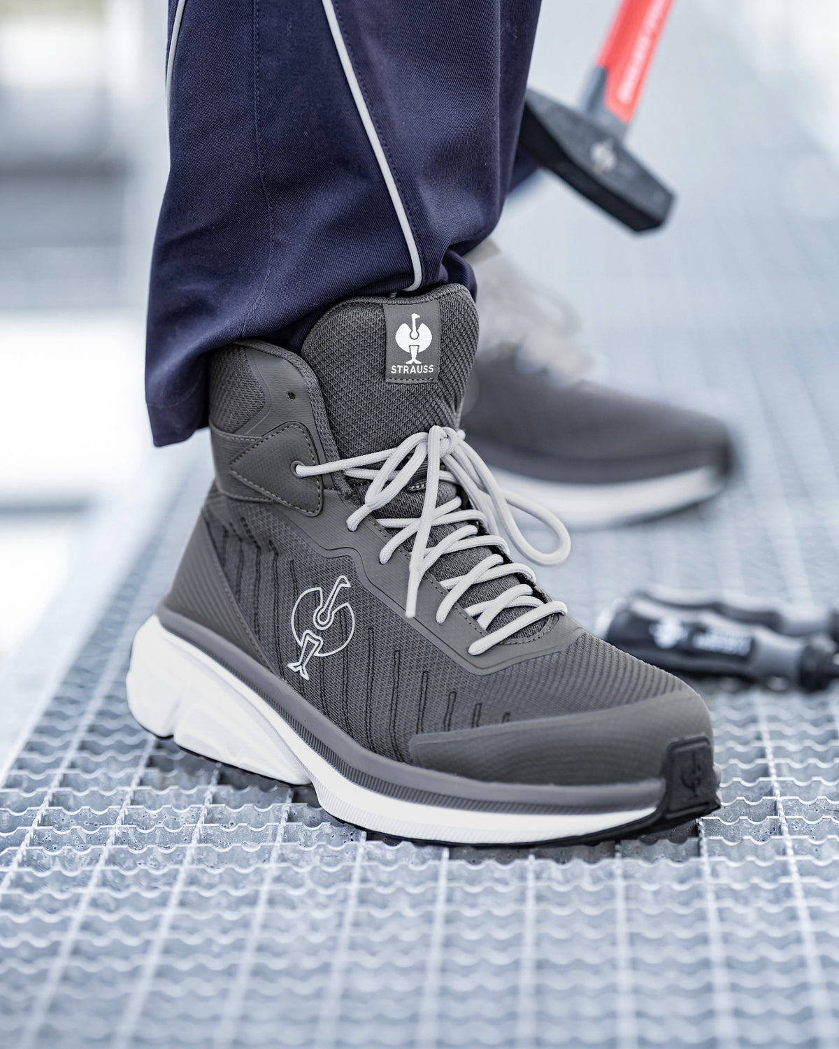 A close-up of a person wearing a dark gray and white high-top athletic boot with white laces, standing on a textured metal grate. The brand logo on the tongue and side reads 'SAS'.