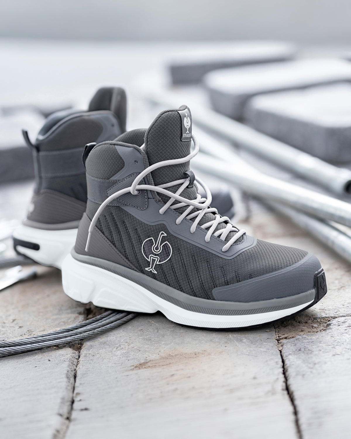 A pair of mid-top athletic sneakers in dark gray and white, featuring a textured mesh upper, white laces, and a white sole, displayed on a weathered wooden surface with a blurred industrial background.