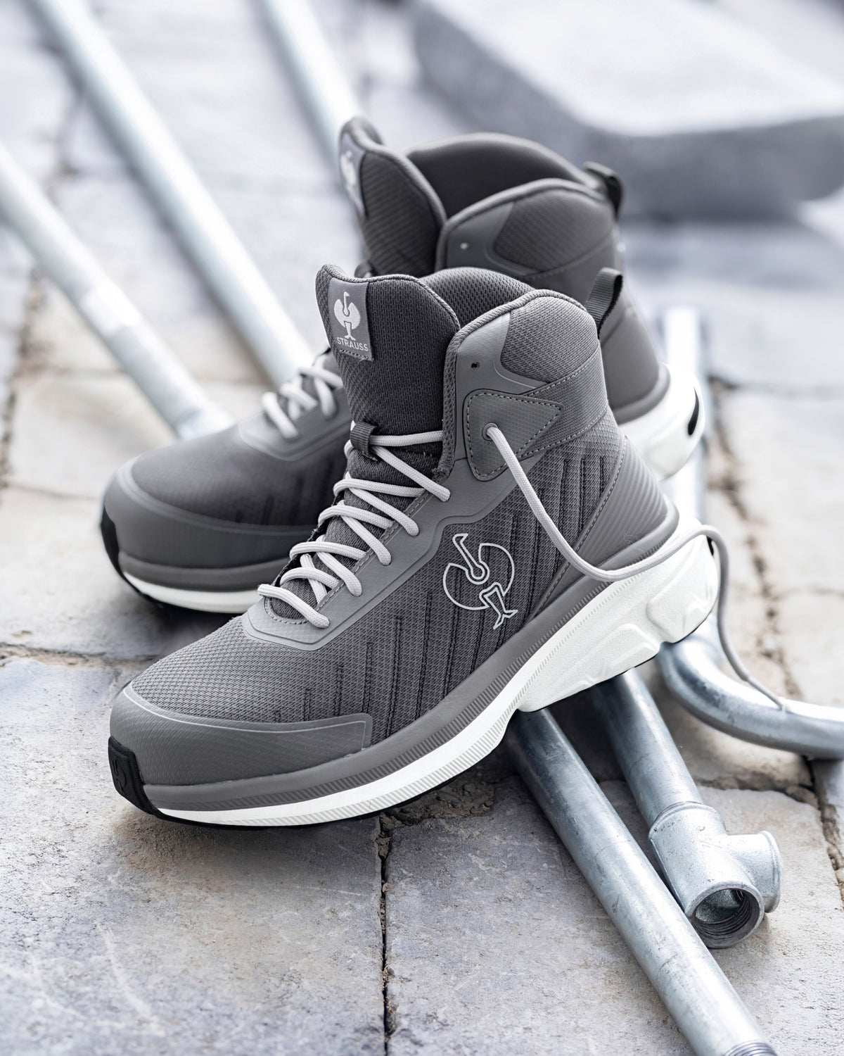 A pair of gray high-top sneakers with white midsoles and a white Spurs logo, resting on metal pipes over a stone pavement background.