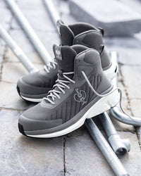 A pair of gray high-top sneakers with white midsoles and a white Spurs logo, resting on metal pipes over a stone pavement background.