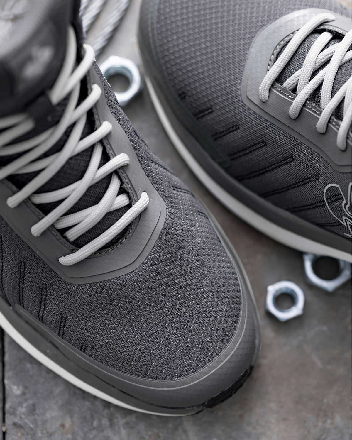 Close-up of a pair of dark gray athletic sneakers with white laces, shown on a textured gray stone surface with small metal hexagonal nuts as props.