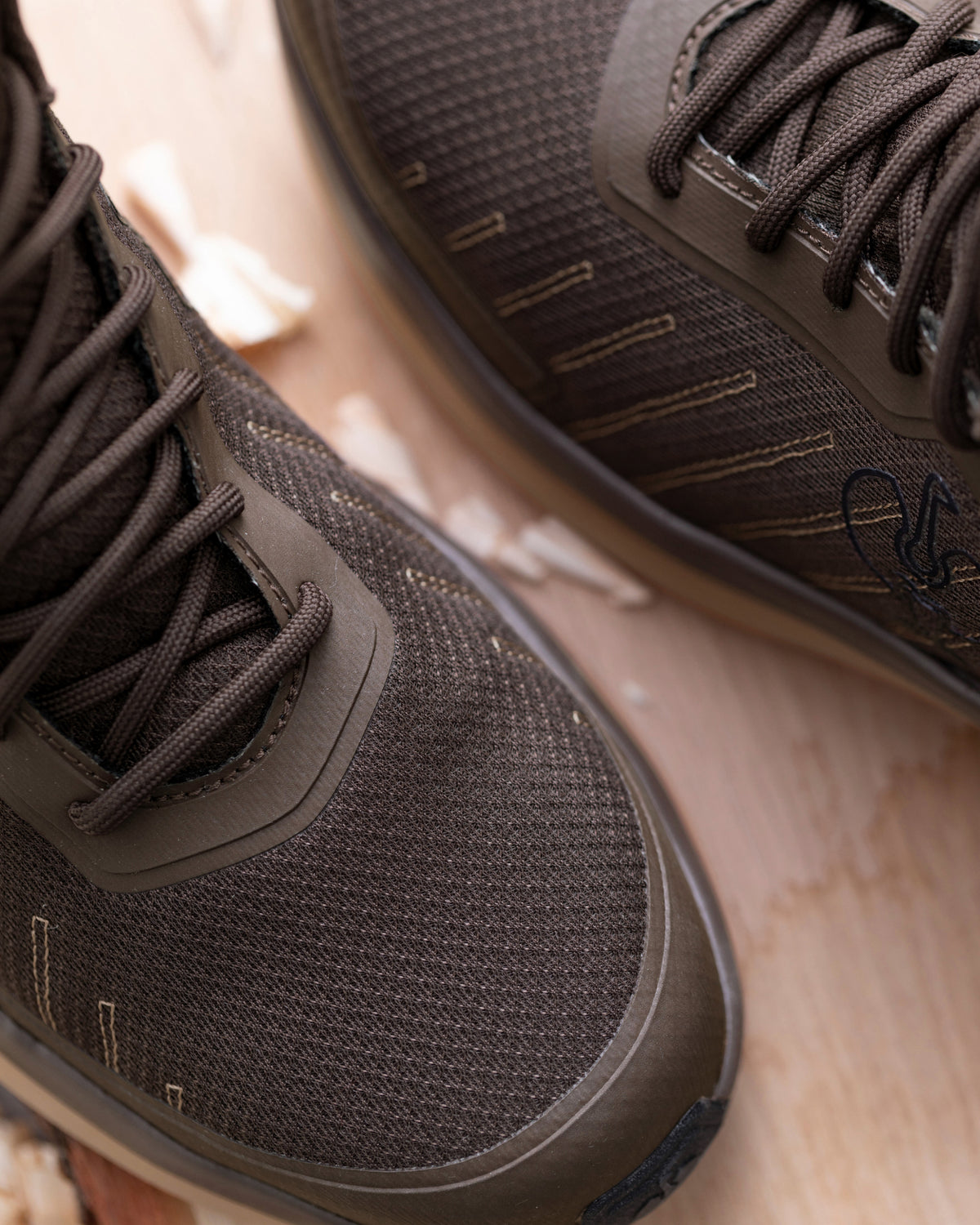 Close-up of a pair of dark brown mesh athletic shoes with matching laces, resting on a light wood surface.