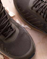 Close-up of a pair of dark brown mesh athletic shoes with matching laces, resting on a light wood surface.