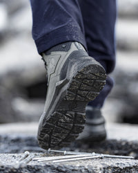 A close-up of a person's foot wearing a gray and black rugged work boot, stepping over metal nails on a rough concrete surface. The person's dark navy pants are also visible.