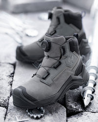 A pair of mid-cut tactical boots in a monochrome gray colorway, displayed on stone tiles with a metal pole as a prop.