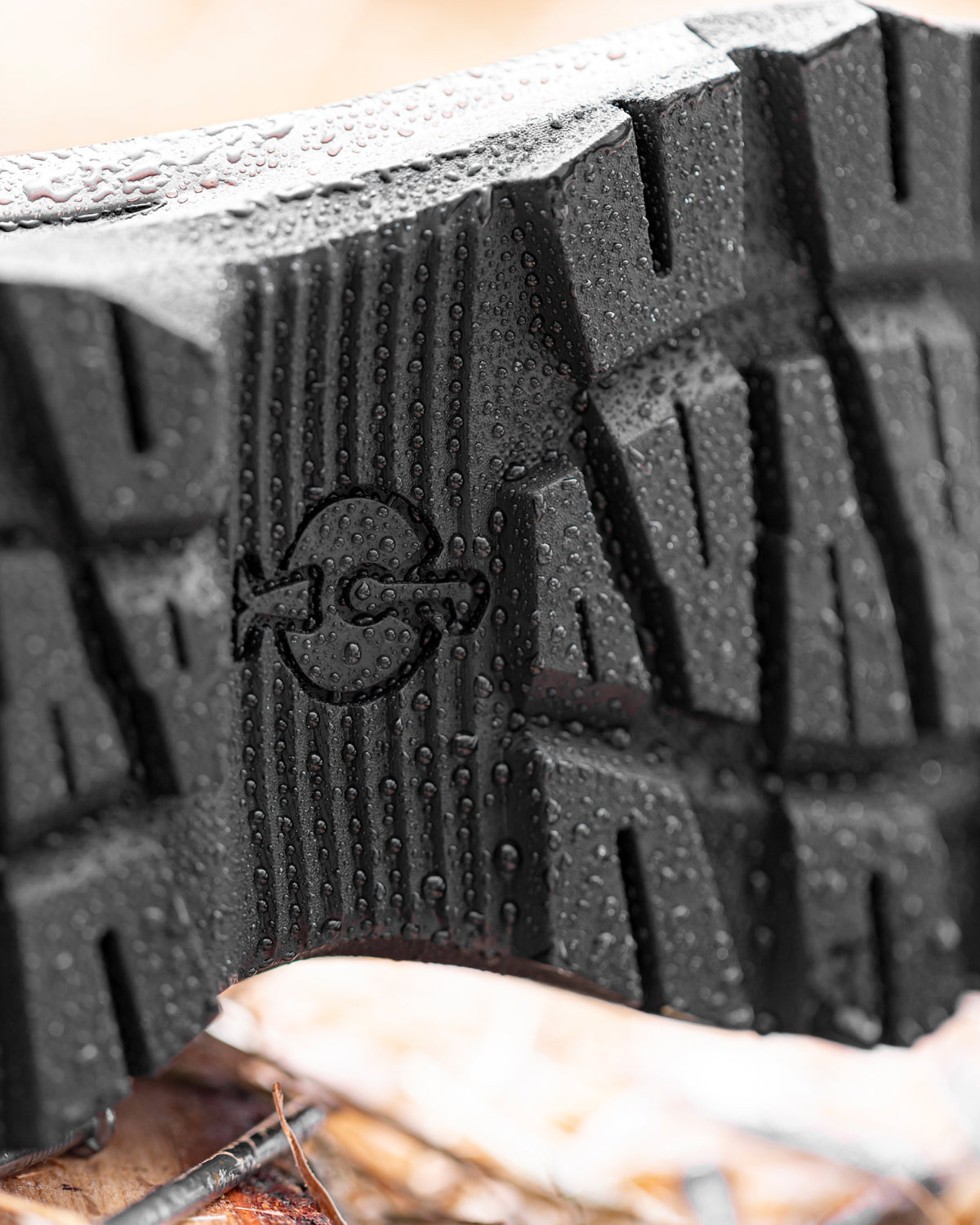 Close-up of a black rubber boot sole with a textured tread pattern and embossed logo, shown outdoors with water droplets on the surface.