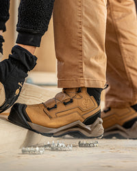 A close-up of a tan work boot with black and gray soles, worn with tan pants and a black work glove. A second pair of work boots and a pair of metal spikes are visible on a concrete surface.
