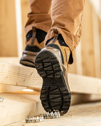 A close-up lifestyle shot of a person wearing tan and black work boots, stepping over a chain of metal spikes on a wooden floor inside a construction site.
