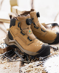 A pair of tan and black BOA work boots with a rugged sole, shown resting on wood shavings and metal nails in a workshop setting.