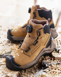 A pair of tan and black work boots with dial-up closure straps, placed on wood shavings with tools in the background.