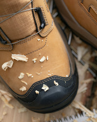 A close-up of a tan leather work boot with a black rubber toe cap, resting on wood shavings and saw blades.