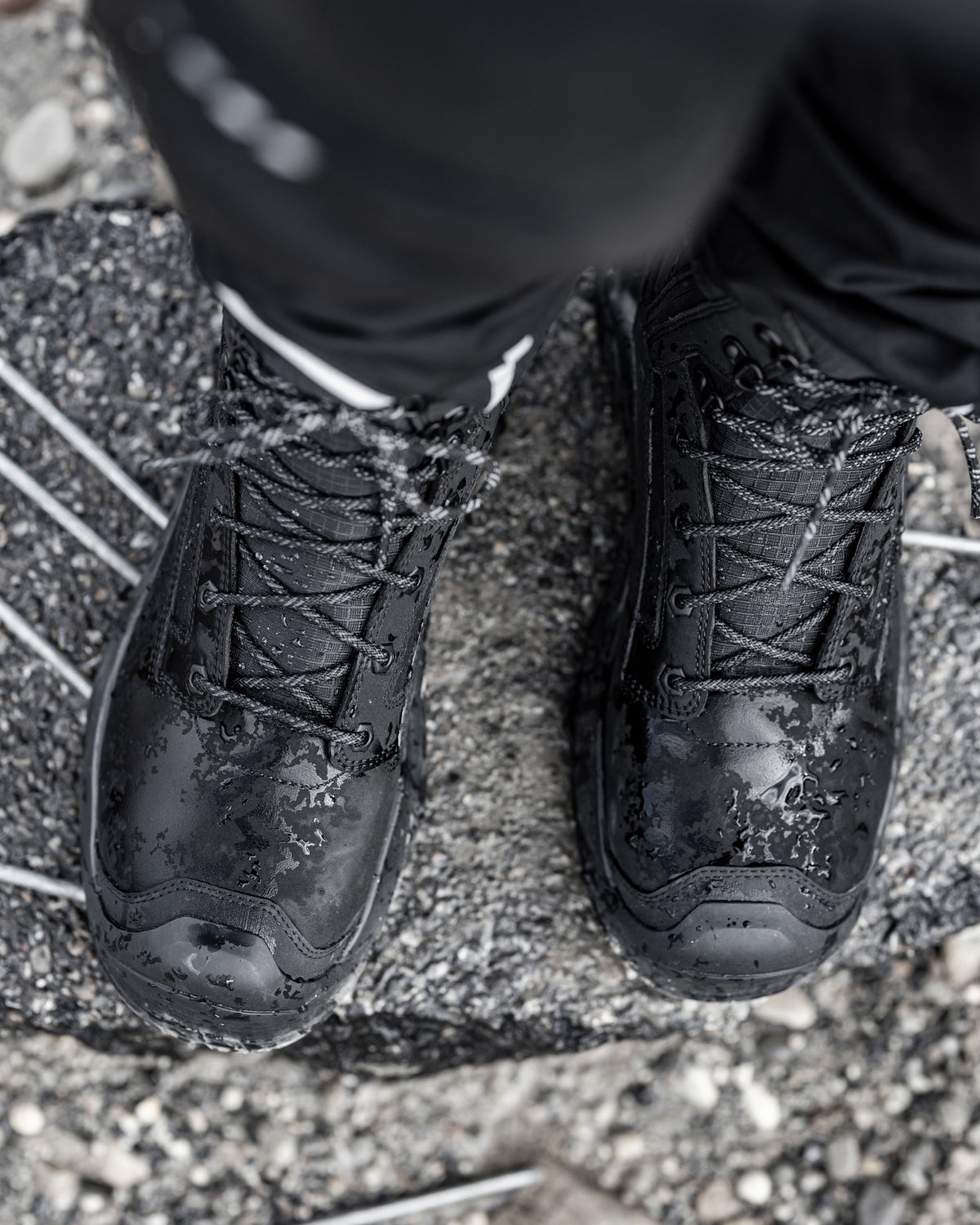 Black waterproof lace-up boots with water droplets on the surface, worn with black pants on a wet gray stone surface.