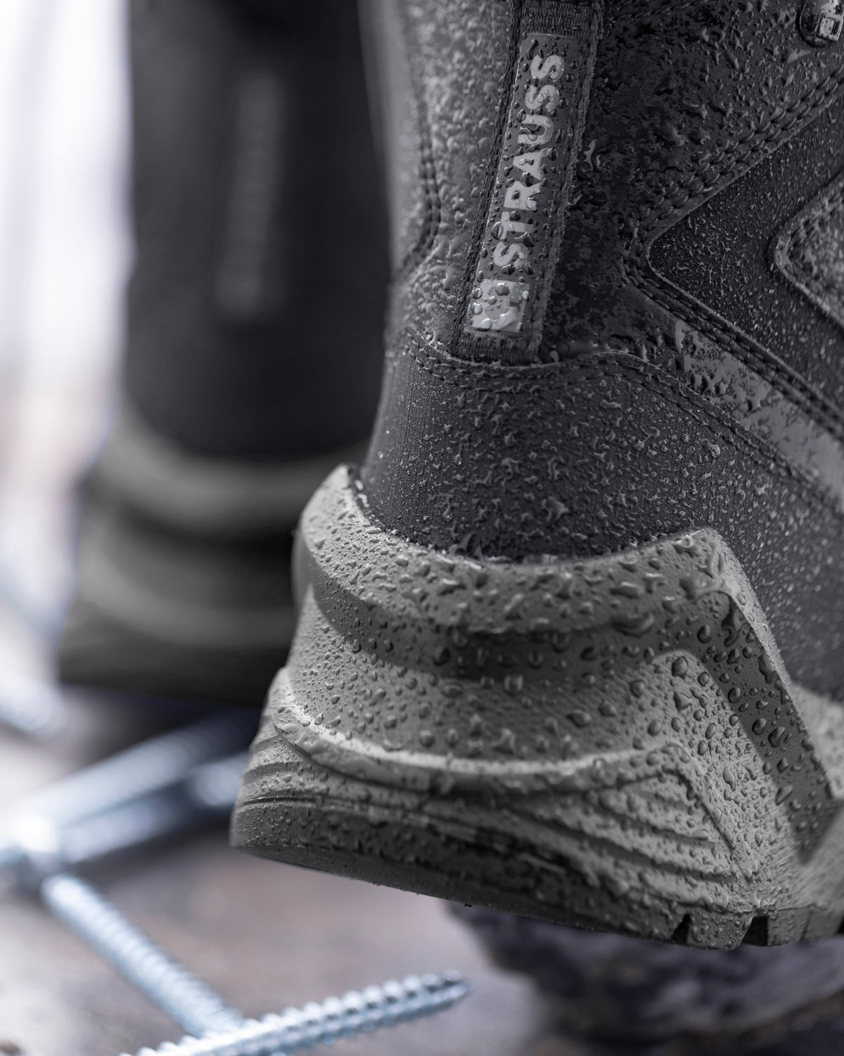 Close-up of a black Draupus Strauss waterproof boot with water droplets on the textured rubber sole and upper, showing the brand label on the heel.