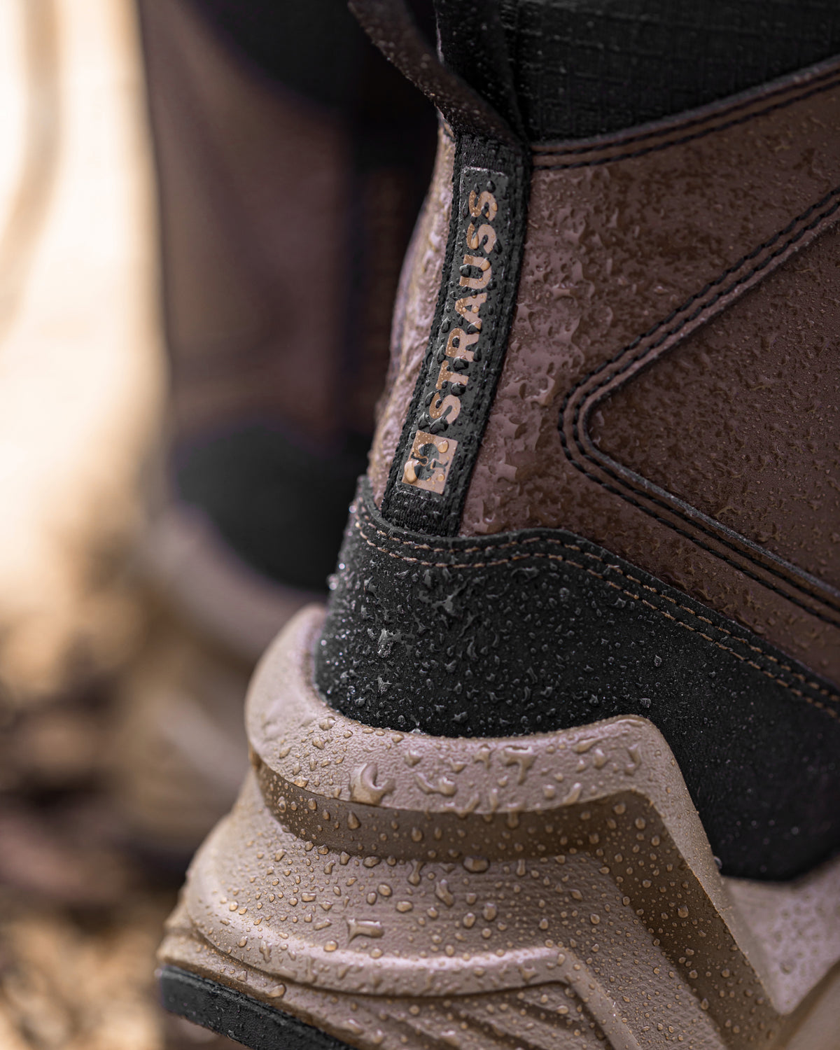 Close-up of a brown and black waterproof boot with water droplets on the surface, showing the 'STRAUSS' brand tab on the heel.