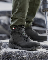A close-up of a person wearing a black leather hiking boot with olive green pants, standing on a stone surface with a metal trekking pole visible.