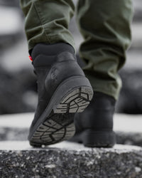 A close-up shot of a person's feet wearing black high-top boots and olive green pants, walking on a wet, dark asphalt surface.