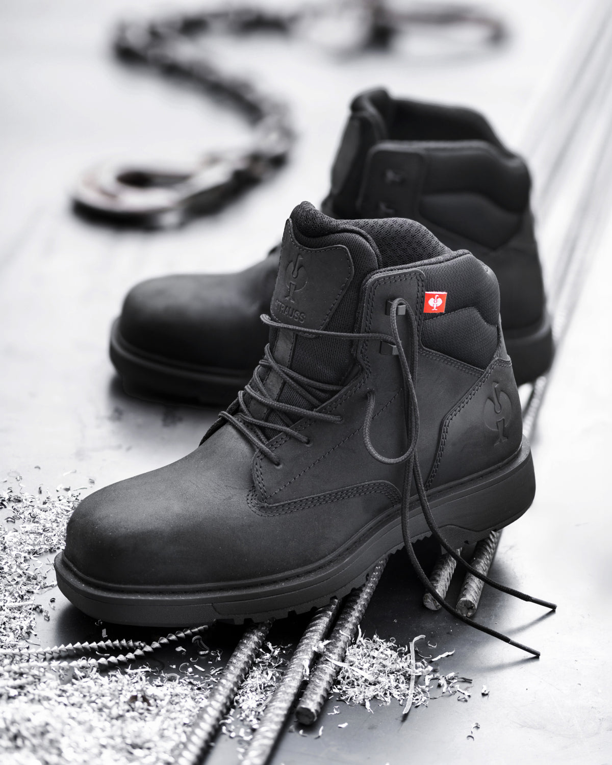 A pair of black high-top work boots arranged on a metallic surface with rebar and small metal shavings. One boot is in sharp focus in the foreground, while the second is slightly blurred behind it.