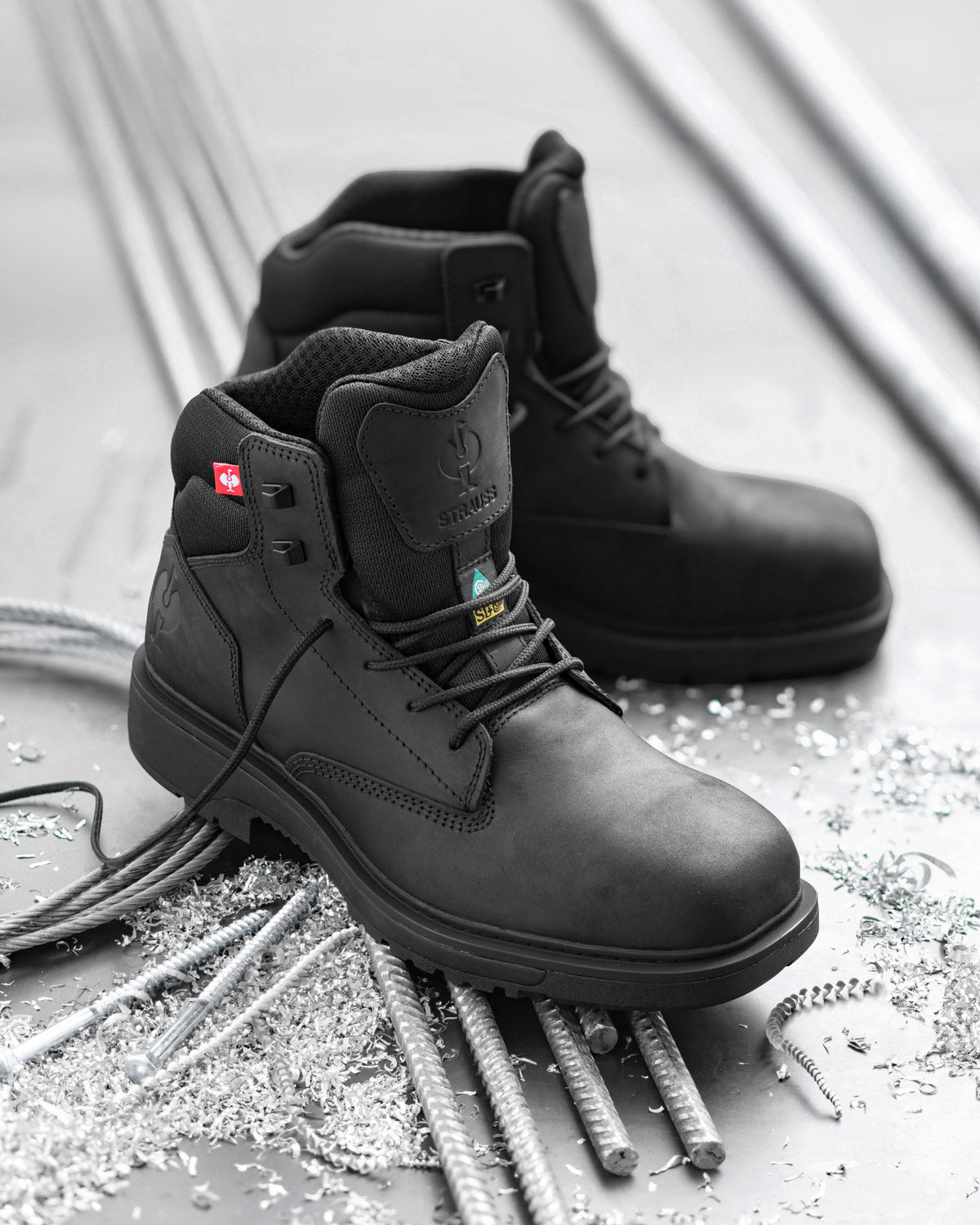 A pair of black industrial safety boots with steel toe protection, displayed on a gray concrete floor surrounded by rebar and metal shavings. The foreground boot is angled to show the side profile, lace-up closure, and heel, while the second boot sits behind it.