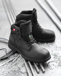 A pair of black industrial safety boots with steel toe protection, displayed on a gray concrete floor surrounded by rebar and metal shavings. The foreground boot is angled to show the side profile, lace-up closure, and heel, while the second boot sits behind it.