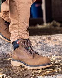 A close-up of a person wearing a pair of brown leather work boots with tan pants, standing on a rustic wooden surface with straw.