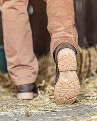 A close-up rear view of a person walking on straw-covered ground, showing tan pants and dark brown boots with the treaded outsole of the right boot in sharp focus.