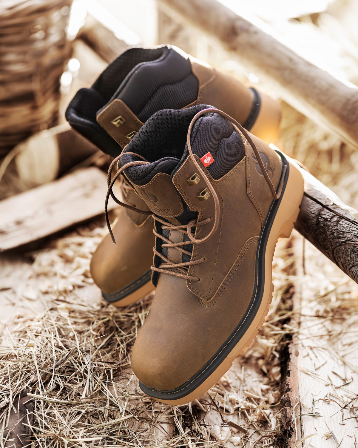 A pair of brown leather lace-up work boots with black lining and tan soles, displayed on a rustic wooden surface with straw and wood logs in the background.