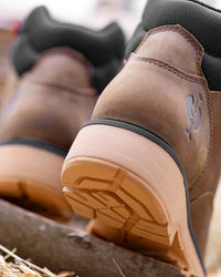 Close-up of a brown leather work boot with a tan rubber sole and black trim, photographed outdoors on a wooden surface with another boot blurred in the background.