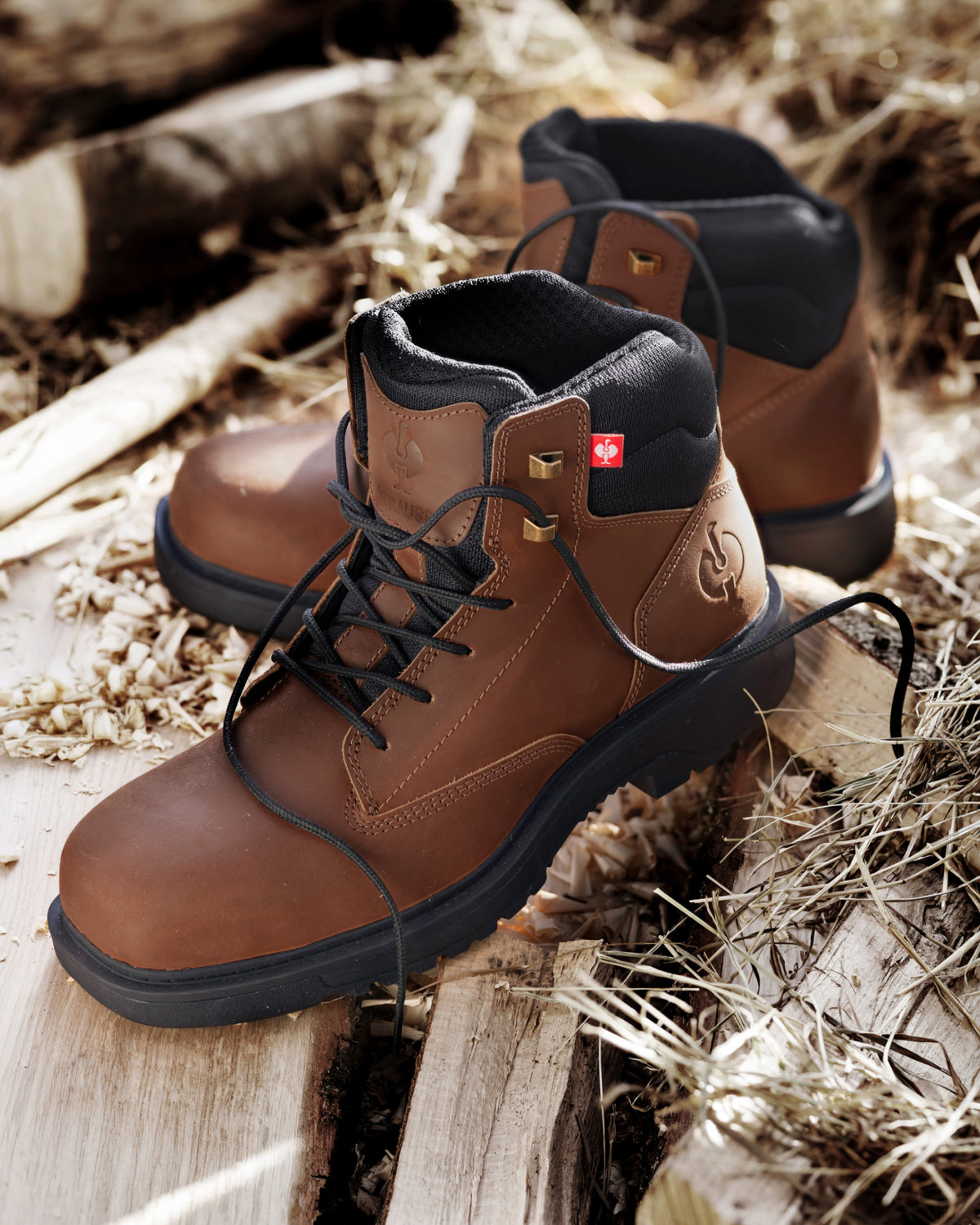 A pair of brown leather lace-up boots with black soles and black lining, placed on weathered wooden planks outdoors with dry grass and logs in the background.