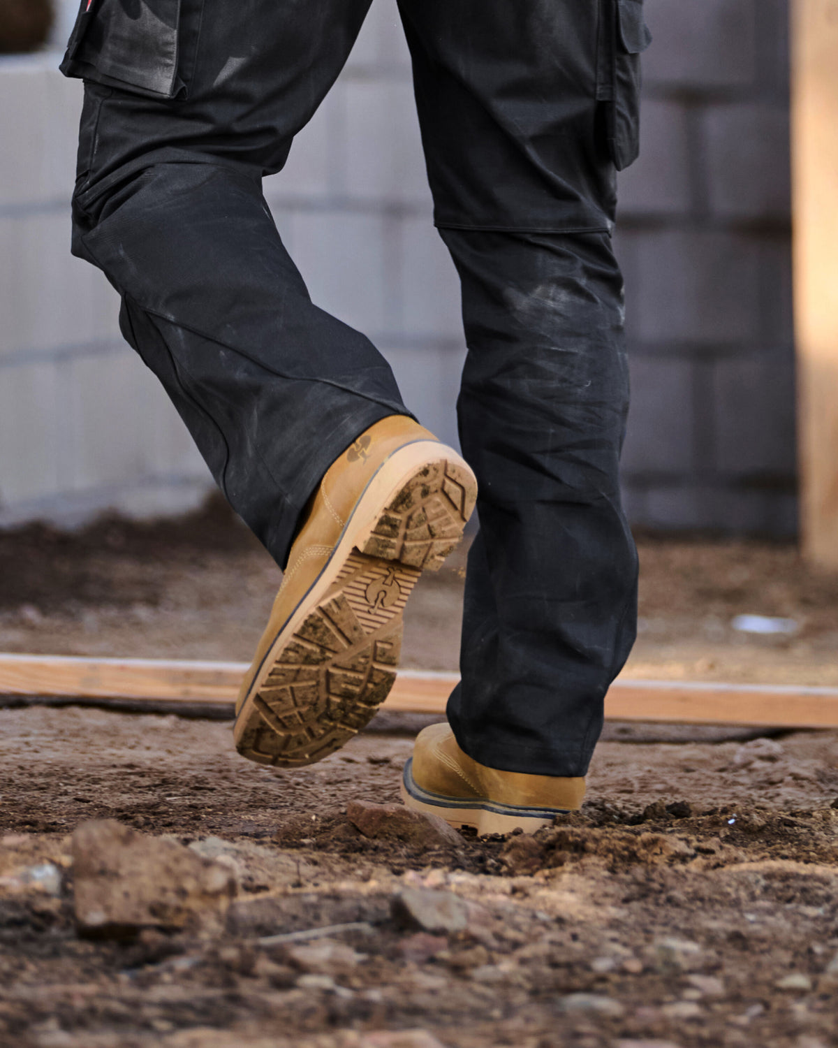 A close-up of a person's legs wearing black work pants and tan work boots, stepping onto a dirt construction site.