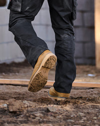 A close-up of a person's legs wearing black work pants and tan work boots, stepping onto a dirt construction site.