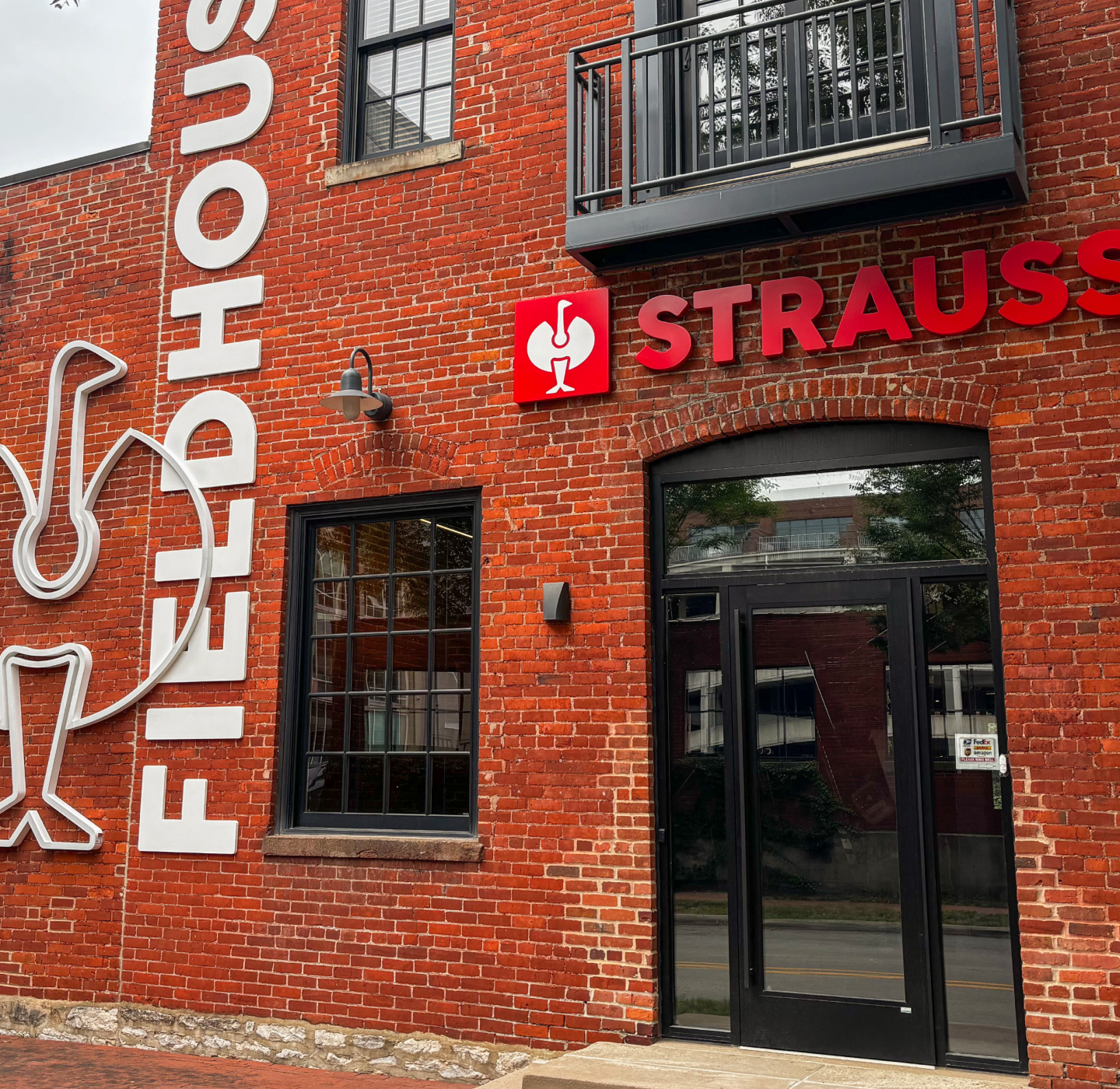 Red brick building with black-framed windows and doors, a balcony, and a sign reading STRAUSS next to a white bird logo. The word FIELDHOUSE is vertically painted on the wall.